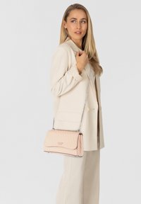 Vrouw in beige pak met een gewatteerde lichtroze schoudertas met kettingband, kijkt zijwaarts tegen een effen lichte achtergrond.