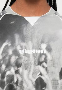 Közelkép egy olyan személyről, aki szürkeárnyalatos sportpólót visel, amelyen tömegnyomás található, és a mellkason középen az "UMBRO" logó látható.