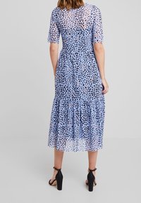 Průhledné modré šaty s puntíkovaným vzorem v černé a oranžové barvě, skládající se z vrstev, s krátkými rukávy a délka po lýtka. Černé vyšší boty.