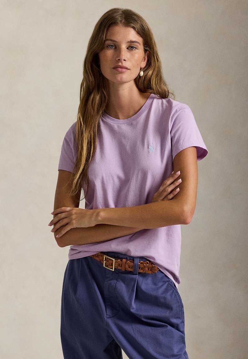 Ανοιχτό μωβ βαμβακερό t-shirt με κοντά μανίκια και στρογγυλή λαιμόκοψη. Διαθέτει ένα μικρό μπλε κεντητό λογότυπο. Σκούρο μπλε παντελόνια και πλεκτή καφέ ζώνη.