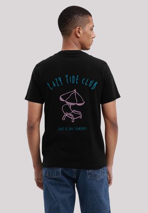 Mann trägt ein schwarzes T-Shirt mit blauem und pinkfarbenem Text und Illustration: "Lazy Tide Club" über einer Zeichnung eines Strandstuhls und Sonnenschirms, trägt blaue Jeans.
