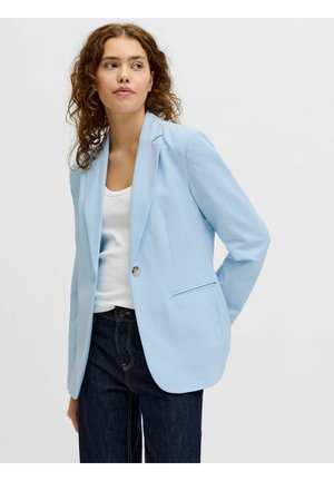 Vrouw met krullend haar draagt lichtblauw colbert, wit topje en donkere spijkerbroek, staat met handen achter de rug en kijkt iets opzij.