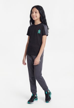 Tüdruk seisab, kannab musta ja halli spordiriietust türkiissiniste detailidega ning musti tossusid türkiissiniste detailidega, naeratab ja vaatab paremale.