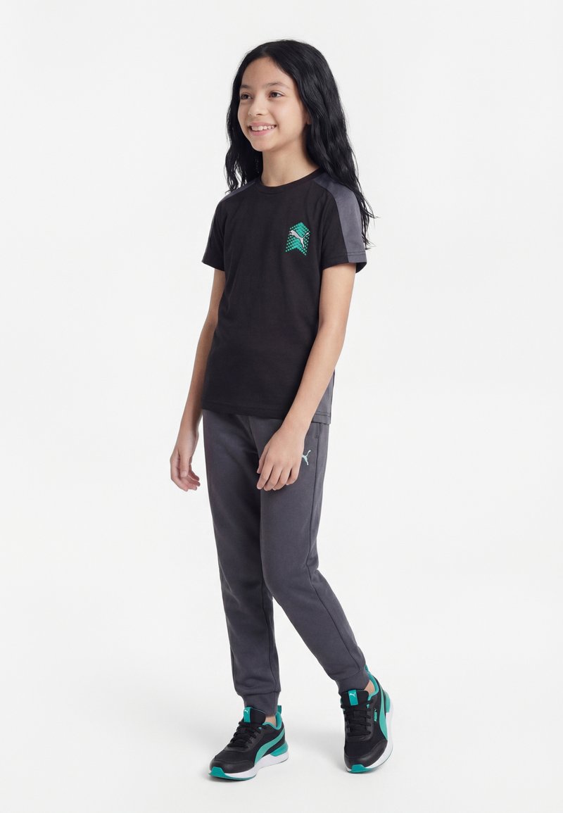 Fille debout, portant une tenue de sport noire et grise avec des accents bleu sarcelle et des baskets noires avec des détails bleu sarcelle, souriante et regardant vers la droite.