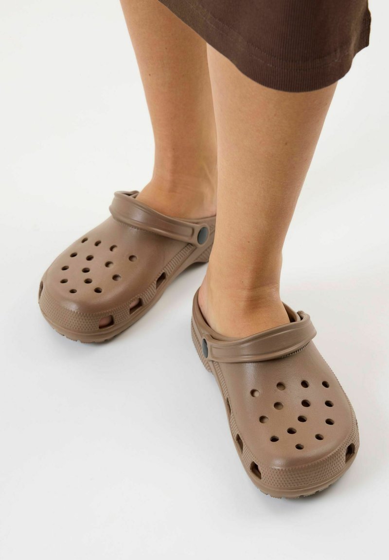 Personne portant des sabots marron à enfiler avec des trous de ventilation et des sangles arrière, debout sur un sol blanc.