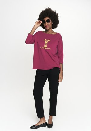 Burgundowa bluzka z długim rękawem z napisem "LUŹNE ROZMOWY ... SĄ DLA PTAKÓW", zestawiona z czarnymi spodniami i czarnymi balerinami, pozująca w okularach przeciwsłonecznych.