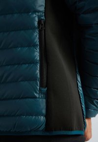 Veste rembourrée avec un extérieur bleu sarcelle foncé, un panneau latéral noir et une poche zippée. Texture lisse avec un motif matelassé.