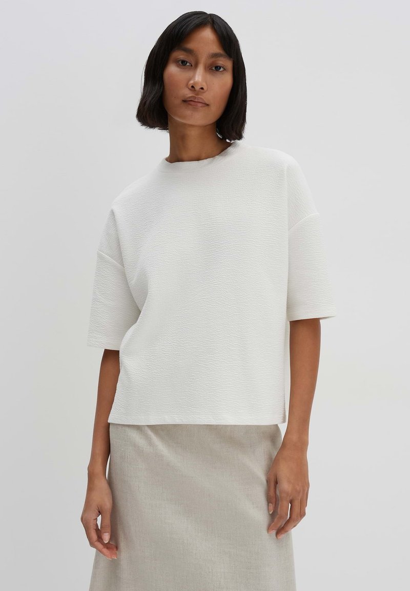 Haut blanc texturé à manches courtes avec un col rond, associé à une jupe gris clair. Tissu lisse, coupe ample et design minimaliste.