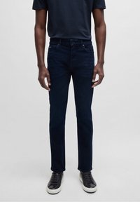 Man med mörkblå slim-fit jeans, svarta sneakers med vita sulor och en marinblå t-shirt, som står mot en ljus, enkel bakgrund.