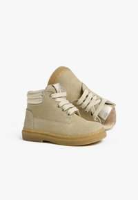 Beige Wildleder-Stiefeletten mit strukturierter Oberfläche, runden Schnürsenkeln, Gummisohle und metallischen Akzenten an der Ferse.