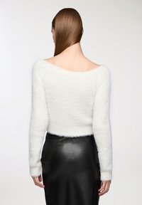 Maglione bianco, morbido e peloso, con spalle scoperte e maniche lunghe, caratterizzato da una texture soffice e un design corto, abbinato a una gonna nera attillata.