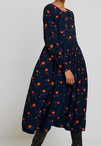 Granatowa sukienka w kwiaty z długimi rękawami, z pomarańczowymi i niebieskimi kwiatami, okrągłym dekoltem oraz marszczoną talią, co tworzy zwiewny kształt.