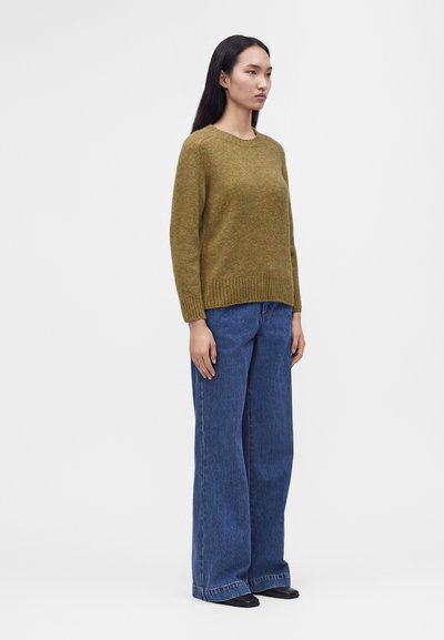 Femme debout portant un pull vert olive, un jean bleu à jambes larges et des chaussures noires sur un fond gris uni.