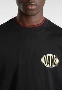 Camiseta de algodón negra con un prominente logo ovalado que dice "VANS" en letras negras en negrita con borde amarillo en el pecho. Diseño de cuello redondo.