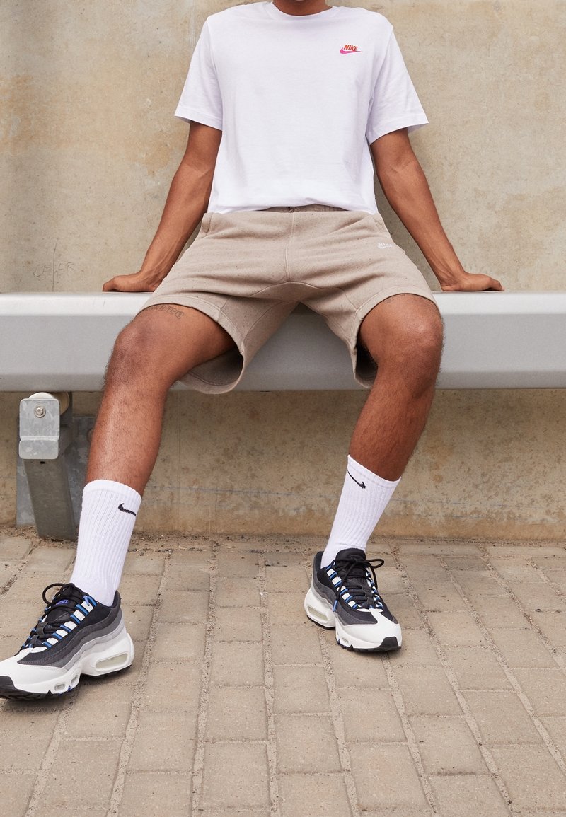 Personne assise sur un banc gris clair portant un t-shirt blanc Nike, un short beige, des chaussettes blanches Nike et des baskets noires et blanches sur une surface pavée.