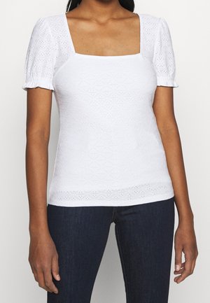 Vrouw draagt een witte blouse met oogjestextuur, opgeblazen korte mouwen en een vierkante halslijn, gecombineerd met donkere jeans.