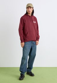 Burgundy hoodie with a front pocket and logo, paired with light blue jeans and black platform shoes, set on a green floor.