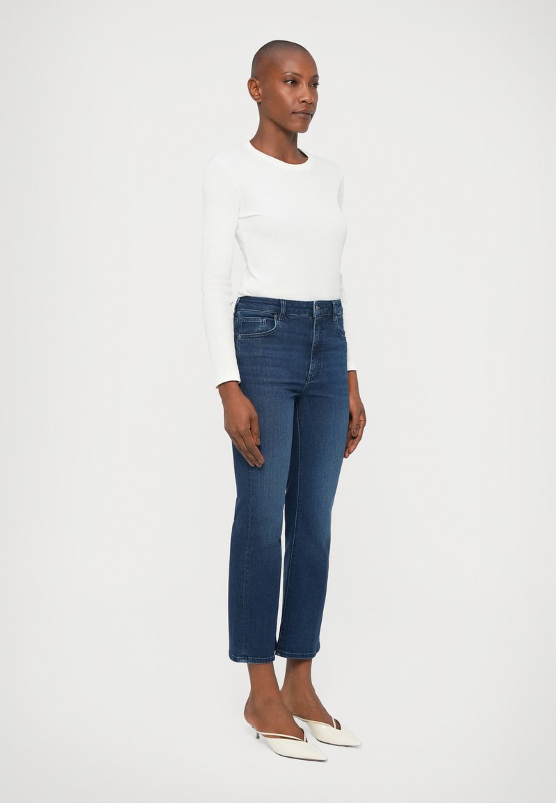 High-waisted blue denim jeans with a slim fit, slight stretch, five-pocket design, and cut-off hem paired with white low-heeled shoes.
