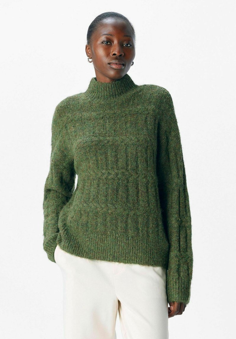 Gebreide groene trui met hoge kraag, gestructureerd patroon en geribde manchetten, gecombineerd met lichtgekleurde broek. Zacht, warm materiaal.