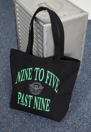 Sac fourre-tout en toile noire avec texte vert indiquant "NEUF HEURES CINQ MINUTES PASSÉES NEUF HEURES." Comprend des poignées doubles et un logo circulaire avec des ailes.