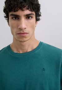 Tricou din bumbac teal cu guler rotund și detaliu subtil al logo-ului. Textură netedă, design minimalist și margine interioară a gulerului în contrast.