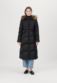 Ilgas juodas pūkinis paltas su dirbtiniu kailio kapu, diržu ant liemens ir siuvinėta tekstūra, derinamas su trumpais mėlynais džinsais ir juodais batais.