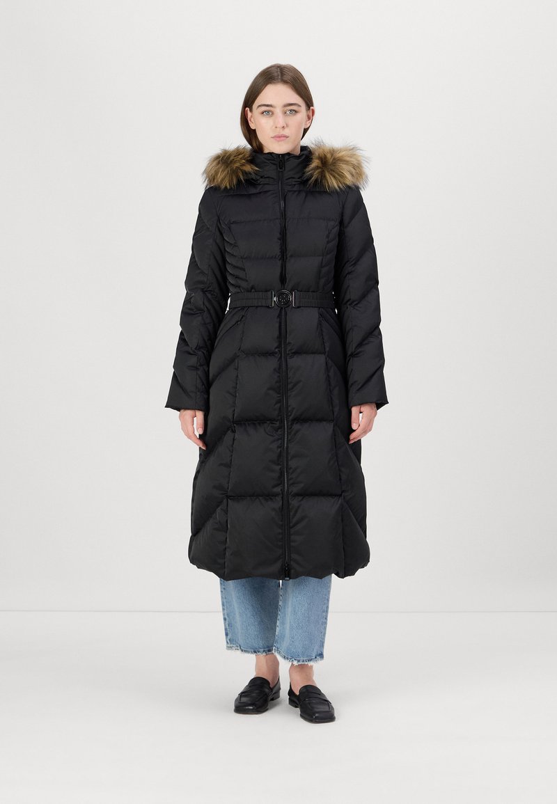 Ilgas juodas pūkinis paltas su dirbtiniu kailio kapu, diržu ant liemens ir siuvinėta tekstūra, derinamas su trumpais mėlynais džinsais ir juodais batais.