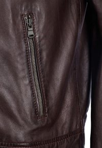 Brun läderjacka med en strukturerad yta, med en dragkedjeficka på vänster sida och kontrasterande sömmar längs fogarna.