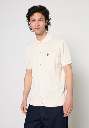 Mann trägt cremefarbenes, kurzärmeliges Polohemd mit kleinem gelben Logo auf der Brusttasche und dunkelblaue Jeans, steht vor weißem Hintergrund.
