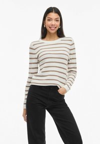 Femme portant un pull rayé blanc et marron et un pantalon noir, souriante avec une main dans la poche, sur fond blanc.