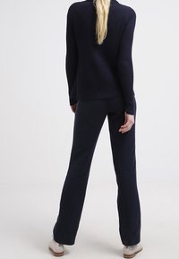 Mujer con cabello largo y rubio, vestida con un suéter negro de canalé, pantalones negros y zapatos blancos, de pie de espaldas contra un fondo liso.