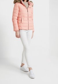 Jasnoróżowa kurtka puchowa z kapturem wykończonym sztucznym futrem, zapinana na zamek, z dwoma bocznymi kieszeniami. Noszona z białymi obcisłymi dżinsami i białymi sneakersami.