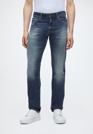 Mand iført blå falmede jeans med lige ben og hvide sneakers, stående med afslappede arme ned langs siden mod en ensfarvet baggrund.