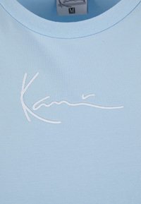 Lyseblå bomulls T-skjorte med ribbestruktur og et hvitt brodert signaturdesign. Lappen viser størrelse medium.