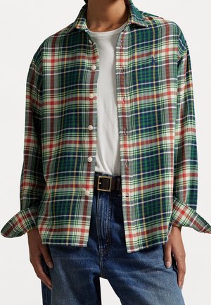 Persoon draagt een blauw, groen en rood geruit overhemd over een wit T-shirt, gecombineerd met een blauwe spijkerbroek en een zwarte riem met een gouden gesp.