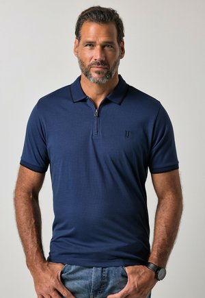 Mand med skæg iført en marineblå kortærmet poloshirt og blå jeans, stående med hænderne nær lommerne mod en ensfarvet baggrund.