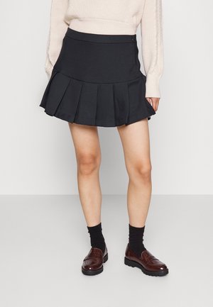 Kvinde iført sort plisseret mininederdel, cremefarvet kabelstrikket sweater, sorte strømper og brune læderloafers, stående på en grå gulv.