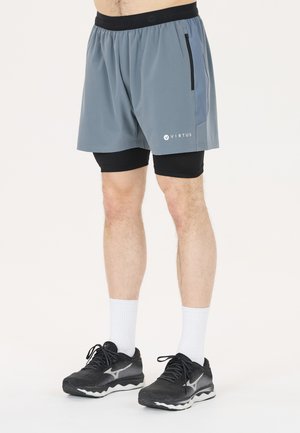 Hombre con pantalones cortos deportivos grises con pantalones cortos interiores de compresión negros, calcetines blancos y zapatillas negras de correr, de pie sobre un fondo liso.