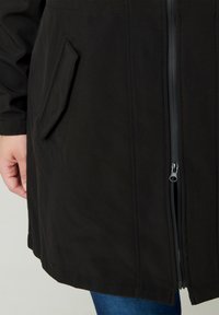 Veste noire à fermeture éclair avec une texture lisse, une poche latérale et une coupe mi-longue. La fermeture éclair est en métal et se trouve au centre.