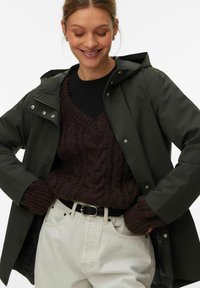 Femme souriante portant une veste à capuche vert foncé par-dessus un pull torsadé marron foncé, un pantalon beige clair et une ceinture noire.