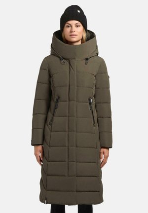 Femme portant un long manteau d'hiver rembourré olive avec un col haut et un bonnet noir, se tenant contre un fond uni.
