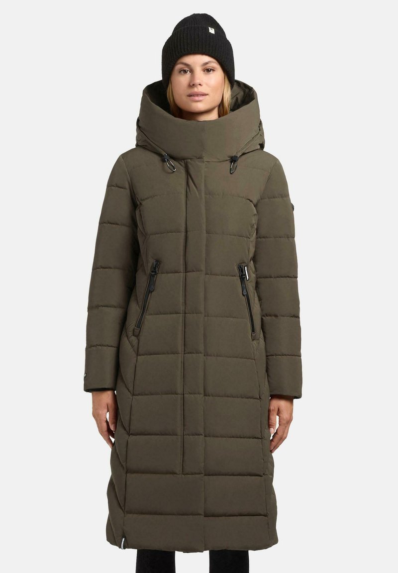 Vrouw in een lange olijfgroene gewatteerde winterjas met een hoge kraag en een zwarte beanie, staande tegen een effen achtergrond.