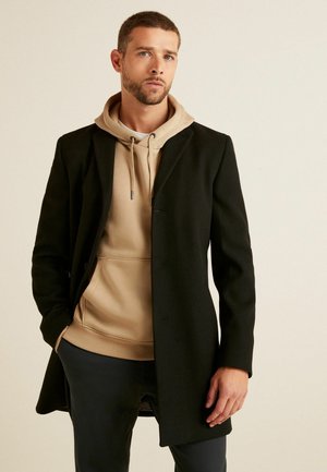Schwarzer Wollmantel über einem beigen Hoodie, der eine Kapuze und einen Kordelzug hat. Der Mantel hat einen einzelnen Knopfverschluss und eine taillierte Passform.