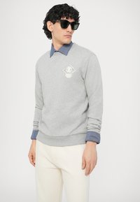 Szara bluza z logo w kształcie diamentu na piersi, nałożona na niebieską koszulę dżinsową. Model nosi ciemne okulary przeciwsłoneczne i jasne spodnie.