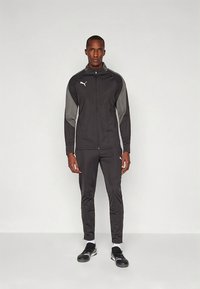 Czarny sportowy dres wykonany z gładkiej tkaniny, z szarymi akcentami na rękawach i bokach. Kurtka na zamek oraz dopasowane spodnie, w połączeniu z czarnymi butami.