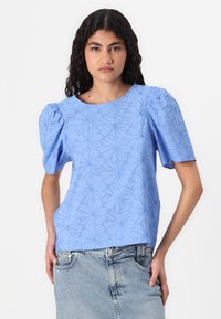 Chemisier bleu clair avec des manches bouffantes, motif floral, col rond, tissu texturé, associé à une jupe en denim.
