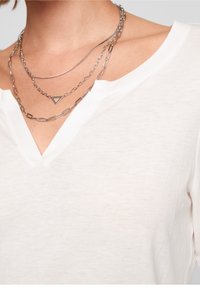 Vit v-ringad blus i mjukt tyg med en slät textur. Den kompletteras av lager av silverkedjor med geometriska inslag.