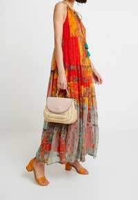 Sukienka maxi z kwiatowym wzorem w czerwonych i pomarańczowych odcieniach, zestawiona z beżową torebką i pomarańczowymi sandałami na obcasie. Lekka, przejrzysta tkanina.