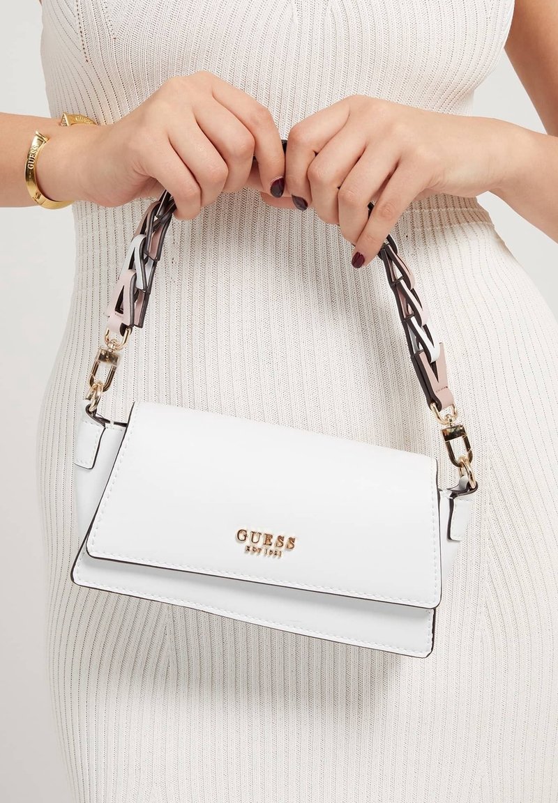 Femme tenant un sac à main blanc Guess avec logo doré et anse chaîne, portant une robe côtelée crème et un bracelet doré.