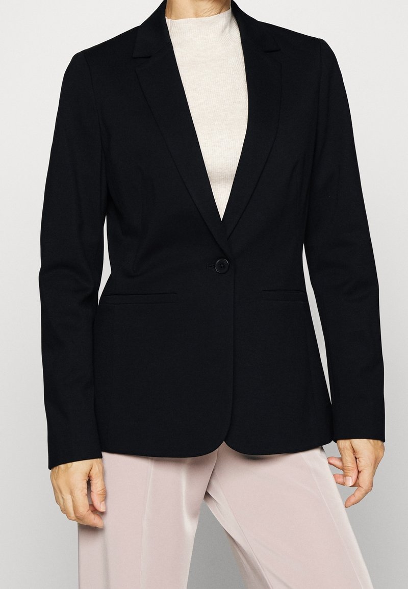 Femme portant un blazer noir à un bouton sur un top blanc et un pantalon beige clair sur un fond uni.
